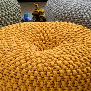May include: A close-up of a yellow knitted pouf or ottoman. The pouf is made of thick, chunky yarn and has a round, slightly indented top.