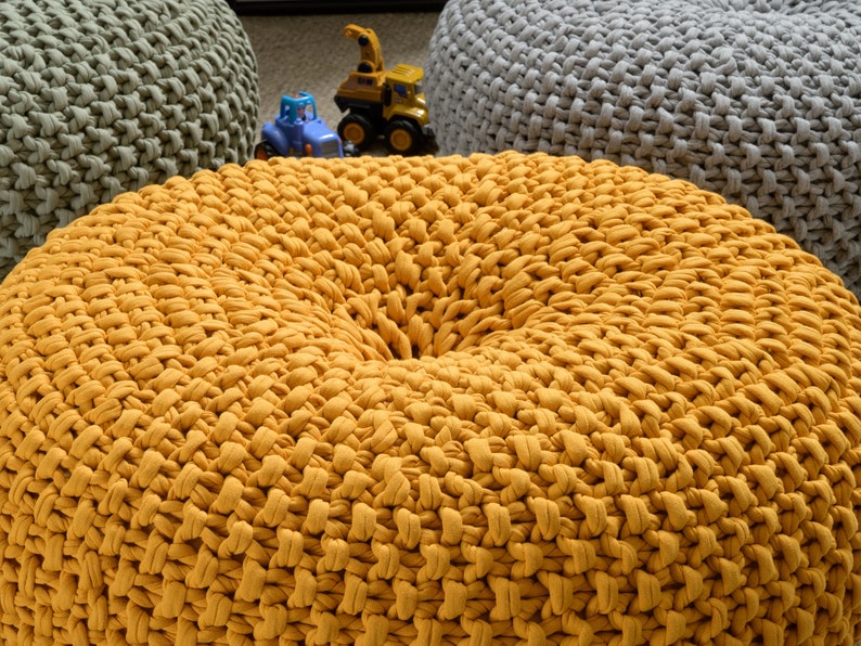 May include: A close-up of a yellow knitted pouf or ottoman. The pouf is made of thick, chunky yarn and has a round, slightly indented top.