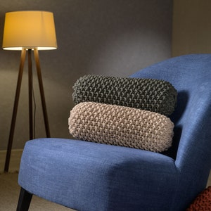 Puede incluir: Tres cojines de sofá de punto en tonos beige, gris y marrón están apilados en un sillón azul. Los cojines están hechos de un tejido de punto grueso y tienen una superficie texturizada.