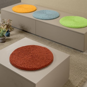 May include: Four round crocheted cushions in different colors: orange, blue, green, and red. The cushions are placed on a light gray bench with drawers.