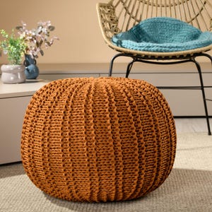 May include: An orange, round knitted pouf with a textured surface sits on a neutral-colored rug. A woven chair with a teal cushion and a small table with vases are in the background. The pouf is a decorative home furnishing.