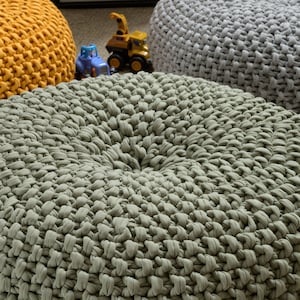 May include: Three round knitted poufs in different colors: mustard yellow, sage green, and light gray. The poufs are made of thick, chunky yarn and have a textured surface.