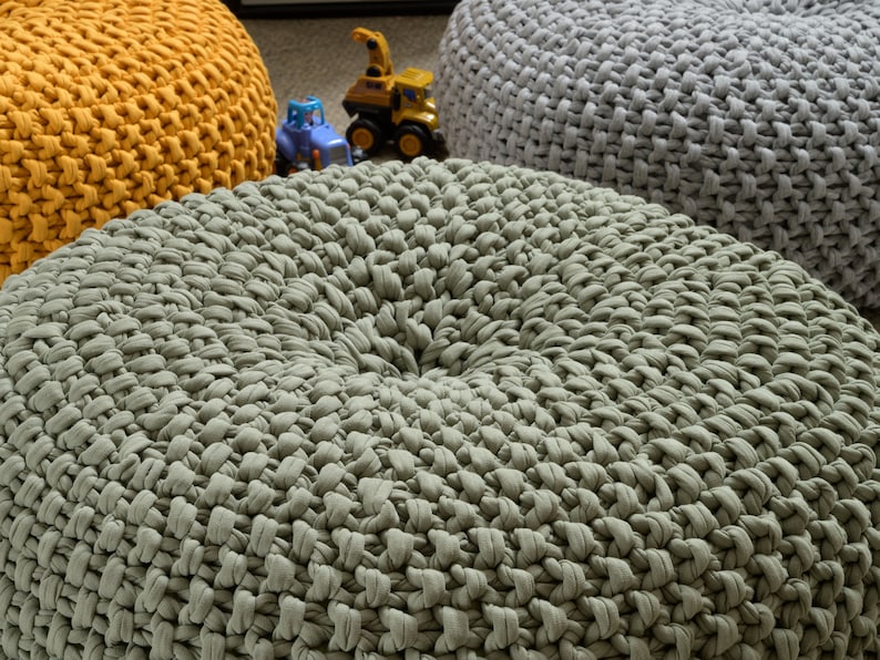 May include: Three round knitted poufs in different colors: mustard yellow, sage green, and light gray. The poufs are made of thick, chunky yarn and have a textured surface.