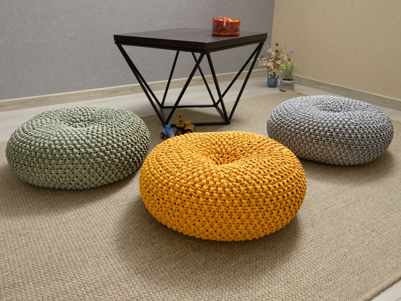 May include: Three round knitted floor cushions in different colors. The cushions are light green, yellow, and gray. The cushions are on a light brown rug.