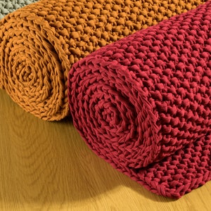 May include: Three rolled up knitted rugs in different colours: grey, orange, and red. The rugs are made of a thick, chunky yarn and have a textured surface.