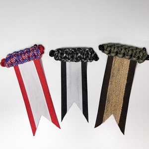 May include: Three fabric ribbon bookmarks with different colored braided tops and white ribbon tails. The bookmarks are red, white, and black, and brown and tan.