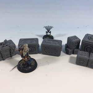 May include: A set of six gray 3D printed miniature boxes with intricate designs. The boxes are arranged in a variety of configurations, including a stack of three and a pair of two. A small gray miniature figure is standing in front of the boxes.