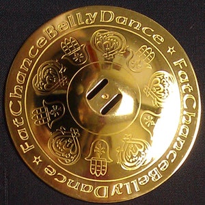 May include: A gold-colored metal belly dance zill with a decorative design featuring the Hamsa hand, a symbol of protection. The zill has two holes for sound and the text "Fat Chance Belly Dance" is engraved around the edge.