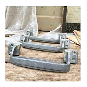 May include: Three silver-toned metal drawer pulls with a matte finish. Each handle features a long, horizontal bar with a slight curve and two rectangular mounting plates at the ends, each containing two pre-drilled screw holes for installation.
