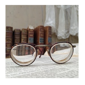 May include: Vintage eyeglasses with a tortoiseshell pattern frame and clear lenses rest on a page of printed text. The glasses have a round shape and are positioned in front of a row of antique books with brown leather covers.