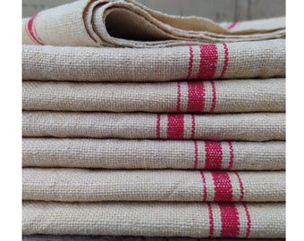 May include: A stack of folded, coarse-textured, off-white fabric cloths. Each cloth features a prominent, vertical, bright red stripe pattern running down the side. The weave of the fabric is visible, showing a thick, rustic, and durable textile construction.