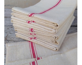 May include: A stack of folded rectangular kitchen towels made of off-white fabric. Each towel features a prominent, vertical red stripe running down the centre. The woven texture of the fabric is visible, showing a consistent pattern throughout the stack.
