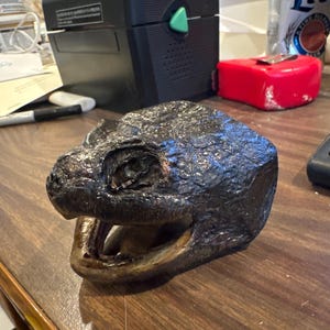 May include: A close-up of a dark brown, textured, turtle head sculpture with an open mouth. The sculpture is made of a hard material and has a realistic appearance.