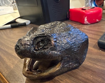 May include: A close-up of a dark brown, textured, turtle head sculpture with an open mouth. The sculpture is made of a hard material and has a realistic appearance.