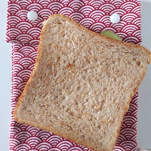 May include: A rectangular fabric sandwich bag featuring a red and white Japanese-inspired wave pattern. The bag is secured with two white plastic snap buttons at the top flap and contains a single slice of toasted whole wheat bread.