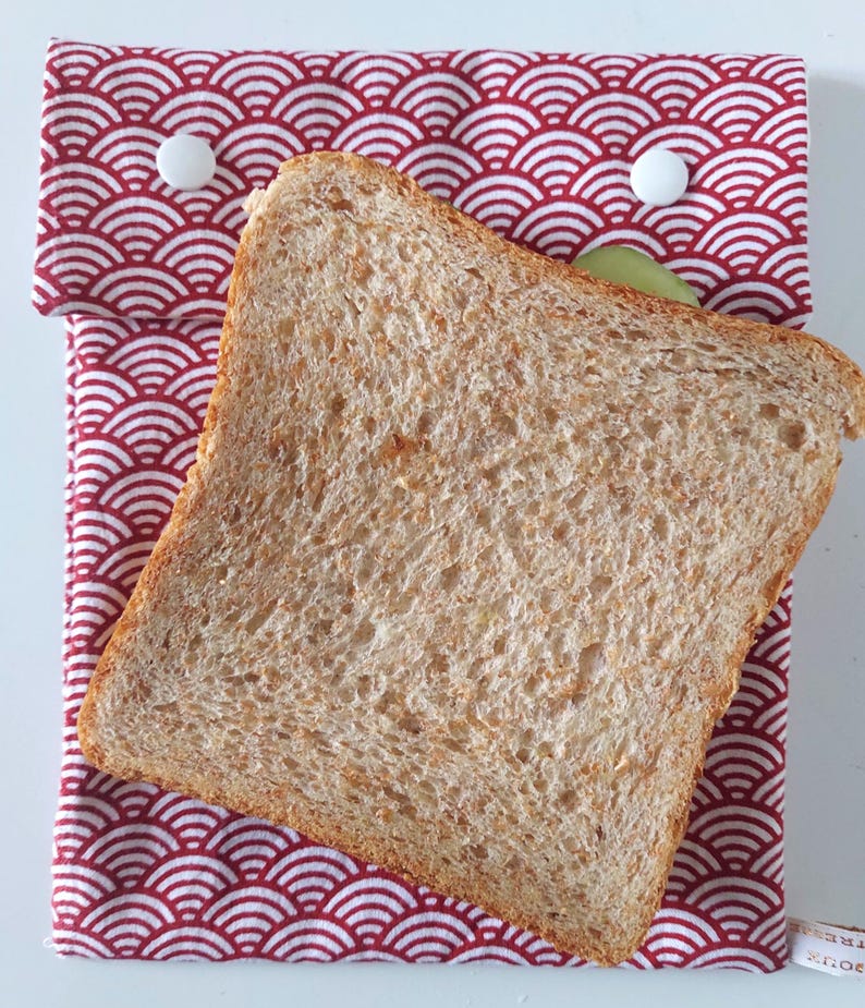 May include: A rectangular fabric sandwich bag featuring a red and white Japanese-inspired wave pattern. The bag is secured with two white plastic snap buttons at the top flap and contains a single slice of toasted whole wheat bread.