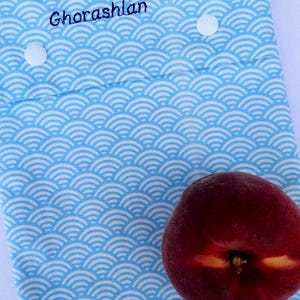 May include: A light blue fabric pouch featuring a white repeating wave pattern, two white snap buttons, and the embroidered name Ghorashlan in dark blue thread. A single fresh peach with red and yellow skin rests on the lower right corner.