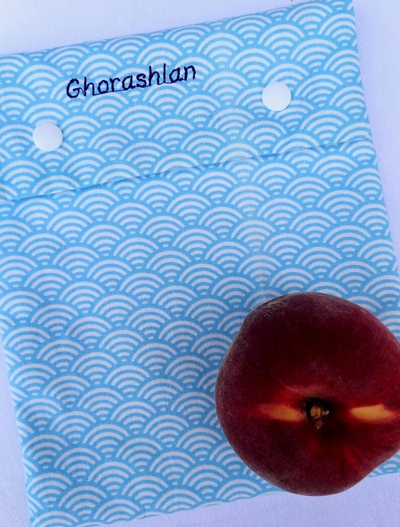 May include: A light blue fabric pouch featuring a white repeating wave pattern, two white snap buttons, and the embroidered name Ghorashlan in dark blue thread. A single fresh peach with red and yellow skin rests on the lower right corner.