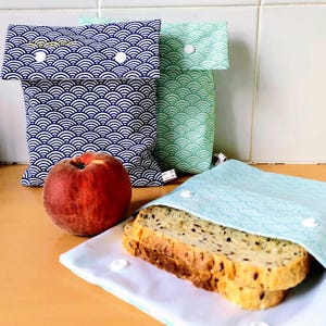 May include: Three fabric snack bags with a repeating wave pattern in dark blue, light green, and light blue. Each bag features a fold-over flap secured with white plastic snaps. A fresh peach sits next to the bags, and a slice of seeded bread rests on the open light blue bag.