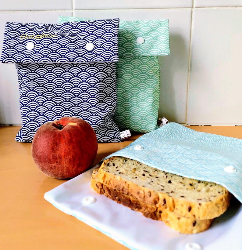 May include: Three fabric snack bags with a repeating wave pattern in dark blue, light green, and light blue. Each bag features a fold-over flap secured with white plastic snaps. A fresh peach sits next to the bags, and a slice of seeded bread rests on the open light blue bag.