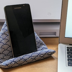 May include: A black Samsung smartphone resting on a triangular fabric stand. The stand features a navy blue and white repeating wave pattern. The device is positioned upright, displaying a dark screen, and is supported by the cushioned fabric base.
