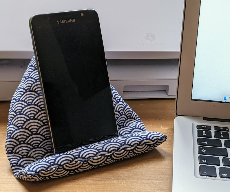 May include: A black Samsung smartphone resting on a triangular fabric stand. The stand features a navy blue and white repeating wave pattern. The device is positioned upright, displaying a dark screen, and is supported by the cushioned fabric base.