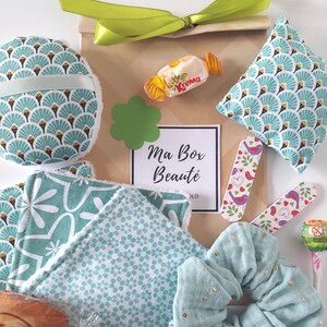 May include: A gift box with beauty products, including a patterned face pad, patterned pillow, scrunchie, and reusable cloths. The box is tied with a green ribbon and contains a candy and a card that reads "Ma Box Beauté".