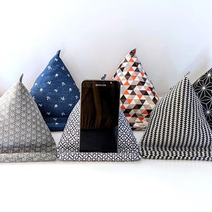 Peut inclure: Plusieurs supports de téléphone triangulaires en tissu, aux motifs et couleurs variés. L'un d'eux maintient un smartphone noir. Les motifs incluent des formes géométriques, des motifs floraux et des imprimés d'oiseaux. La palette de couleurs comprend le bleu, le gris, le noir, le blanc et l'orange.