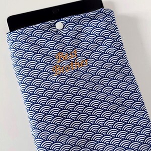 May include: A blue and white patterned tablet sleeve with the words "Best Brother" embroidered in gold. The sleeve is made of fabric and has a button closure. The pattern is a repeating wave design.