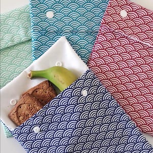 May include: A collection of four fabric pouches with a repeating white wave pattern on mint green, teal, red, and navy blue backgrounds. Each pouch features a white snap closure. One open white pouch contains a banana and two brown square cookies.
