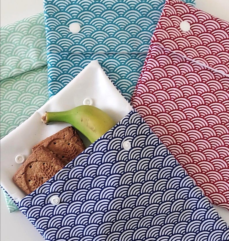 May include: A collection of four fabric pouches with a repeating white wave pattern on mint green, teal, red, and navy blue backgrounds. Each pouch features a white snap closure. One open white pouch contains a banana and two brown square cookies.