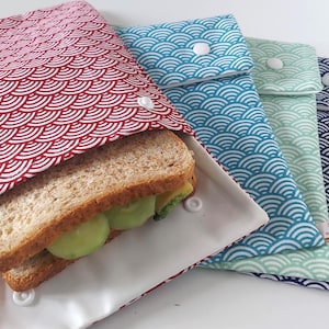 Puede incluir: Envoltorios reutilizables para sándwiches en varios colores, incluyendo rojo, azul y verde, con un patrón de olas. Uno de los envoltorios está abierto, mostrando un sándwich con rodajas de pepino. Los envoltorios tienen cierres de botón a presión blancos.