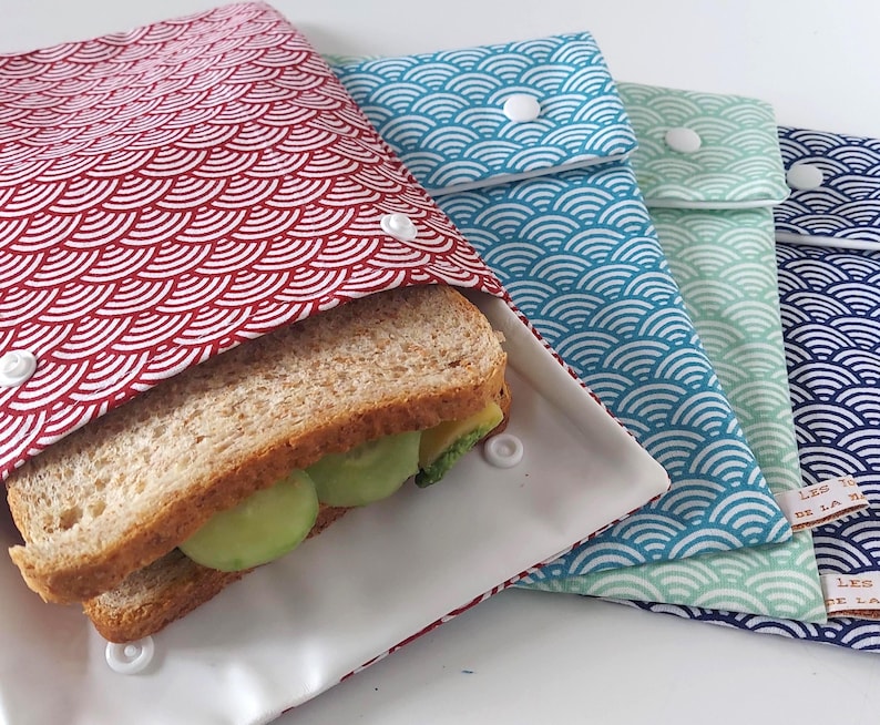 May include: A set of four rectangular fabric sandwich bags featuring a repeating white wave pattern on red, blue, light green, and dark blue backgrounds. Each bag has a white plastic snap closure and contains a sandwich with bread and avocado.