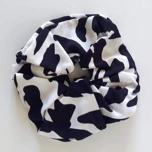 May include: A black and white cow print scrunchie. The fabric is gathered and knotted, forming a voluminous hair accessory. The scrunchie is made of a soft material, suitable for various hairstyles and is a stylish addition.
