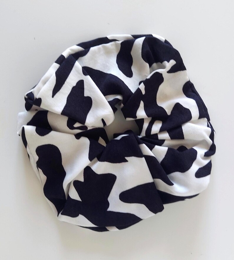 May include: A black and white cow print scrunchie. The fabric is gathered and knotted, forming a voluminous hair accessory. The scrunchie is made of a soft material, suitable for various hairstyles and is a stylish addition.