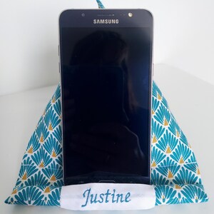 May include: A triangular fabric phone stand featuring a teal and white fan pattern with gold accents. A white horizontal fabric strip at the base is embroidered with the name Justine in teal thread. A black Samsung smartphone rests on the stand.