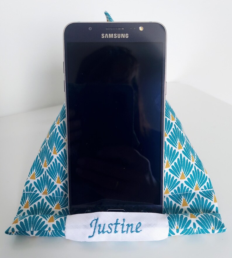 May include: A triangular fabric phone stand featuring a teal and white fan pattern with gold accents. A white horizontal fabric strip at the base is embroidered with the name Justine in teal thread. A black Samsung smartphone rests on the stand.