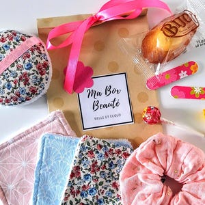 Peut inclure: Un coffret cadeau avec un ruban rose, contenant des produits de beauté. La boîte porte l'inscription "Ma Box Beauté". Contenu : tampons démaquillants à motifs floraux, un chouchou rose et une pâtisserie.