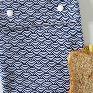 May include: A rectangular fabric pouch featuring a repeating dark blue and white wave pattern. The pouch has a fold-over flap secured by two white circular plastic snap buttons. A slice of whole wheat bread is visible at the bottom right corner.