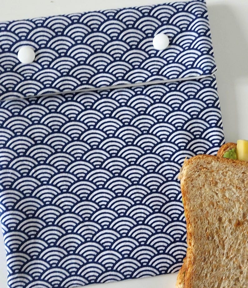 May include: A rectangular fabric pouch featuring a repeating dark blue and white wave pattern. The pouch has a fold-over flap secured by two white circular plastic snap buttons. A slice of whole wheat bread is visible at the bottom right corner.