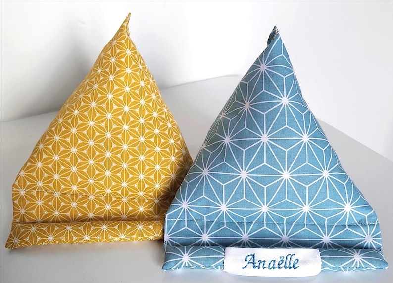 May include: Two triangular fabric phone stands. One is yellow with a white floral pattern, the other is blue with a white geometric design. The blue stand has the name "Ana&euml;lle" embroidered on a white label.