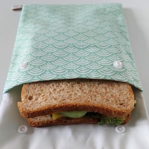 May include: A rectangular fabric sandwich bag featuring a light mint green and white scalloped pattern. The bag is secured with two white plastic snap buttons on the front flap and contains a whole wheat sandwich with visible green and yellow fillings.