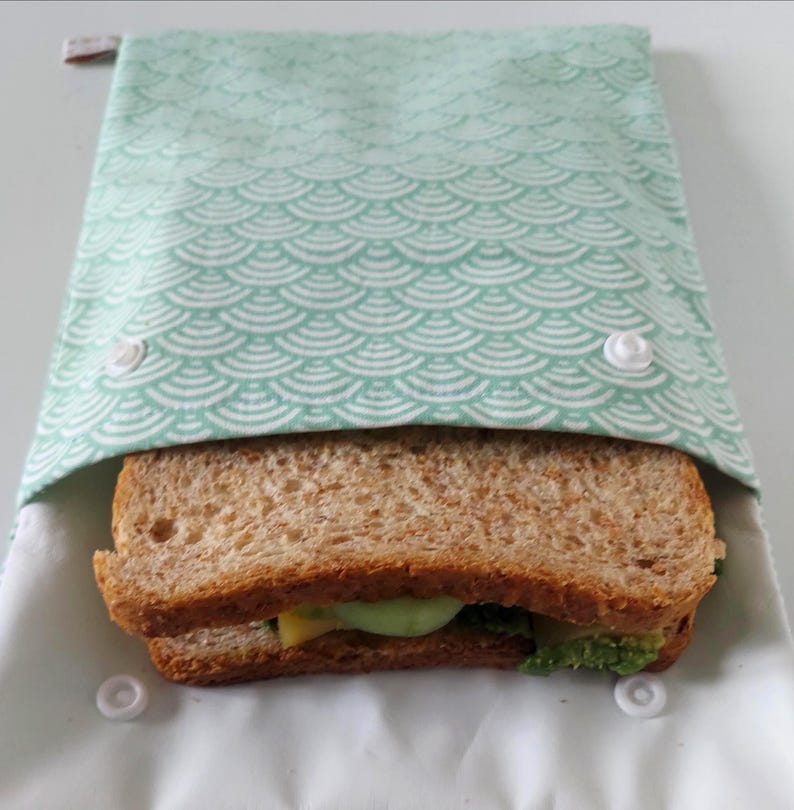 May include: A rectangular fabric sandwich bag featuring a light mint green and white scalloped pattern. The bag is secured with two white plastic snap buttons on the front flap and contains a whole wheat sandwich with visible green and yellow fillings.