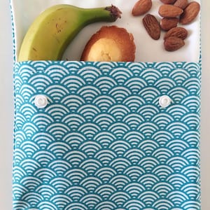 May include: A rectangular fabric snack pouch featuring a teal and white wave pattern with two white plastic snap closures. The pouch is open at the top, revealing a green banana, a golden-brown madeleine cake, and a small pile of brown almonds.
