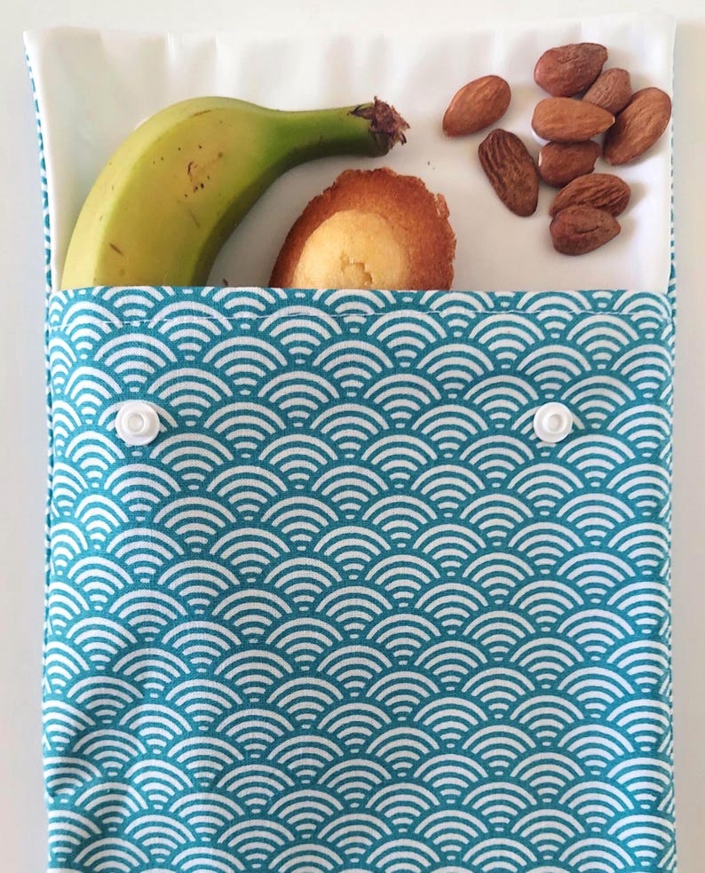 May include: A rectangular fabric snack pouch featuring a teal and white wave pattern with two white plastic snap closures. The pouch is open at the top, revealing a green banana, a golden-brown madeleine cake, and a small pile of brown almonds.