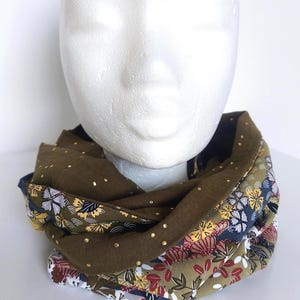 Pode incluir: Cachecol infinito verde-oliva com bolinhas douradas e estampas florais em vermelho, amarelo e azul. O cachecol está enrolado em uma cabeça de manequim branca, mostrando seu design e textura.