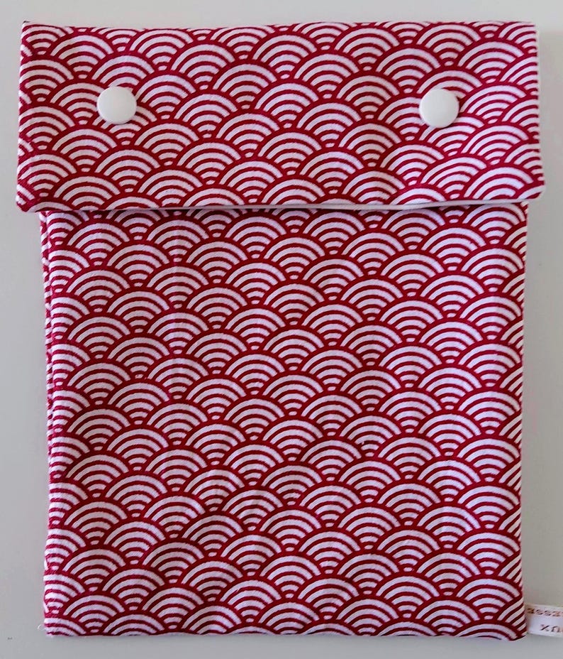May include: A rectangular fabric pouch featuring a repeating red and white wave pattern. The pouch has a fold-over flap secured by two white plastic snap buttons. A small white fabric tag is visible at the bottom right corner of the pouch.