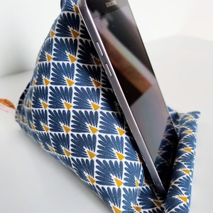 May include: A triangular fabric phone stand featuring a blue and white fan-shaped pattern with small yellow triangles. A black Samsung smartphone is resting against the angled surface of the soft, stuffed textile holder, which is designed to support mobile devices.