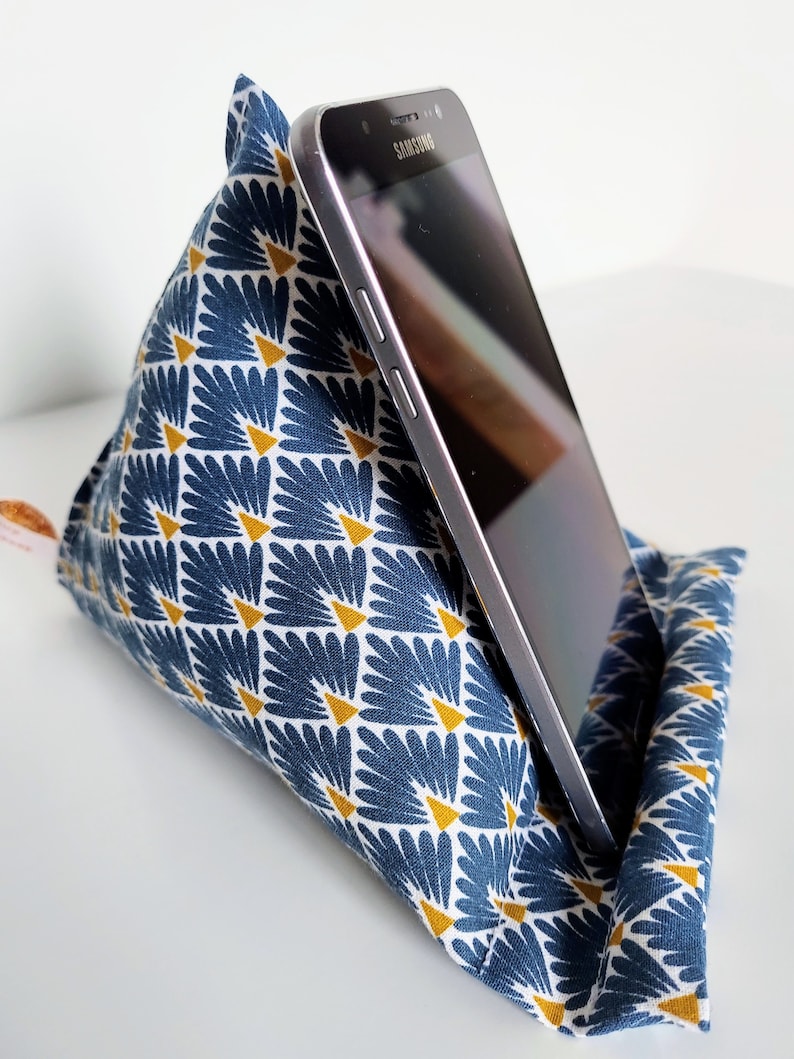 May include: A triangular fabric phone stand featuring a blue and white fan-shaped pattern with small yellow triangles. A black Samsung smartphone is resting against the angled surface of the soft, stuffed textile holder, which is designed to support mobile devices.