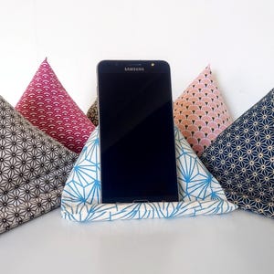 Puede incluir: Un smartphone negro se muestra en un soporte de tela para teléfono. El soporte está sostenido por cuatro cojines triangulares con varios patrones geométricos en tonos de azul, rosa y gris. El soporte está hecho de tela estampada azul y blanca.